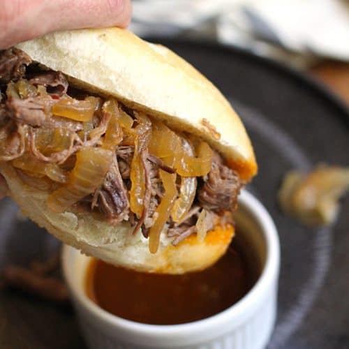 Close-up shot of my hand holding a beef au jus sandwich, dipping it into some au jus in a white bowl, set on top of a black plate.
