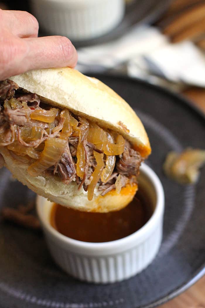 Close-up shot of my hand holding a beef au jus sandwich, dipping it into some au jus in a white bowl, set on top of a black plate.