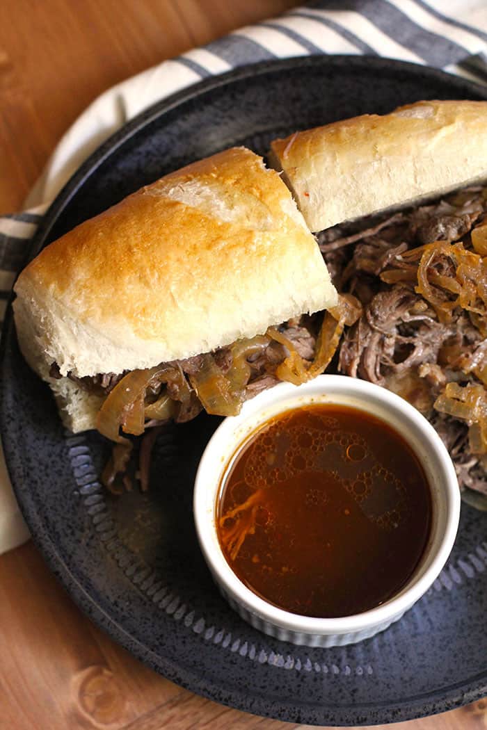 Overhead shot of a beef au jus sandwich, sliced in half, on a black plate with a small white bowl of au jus.