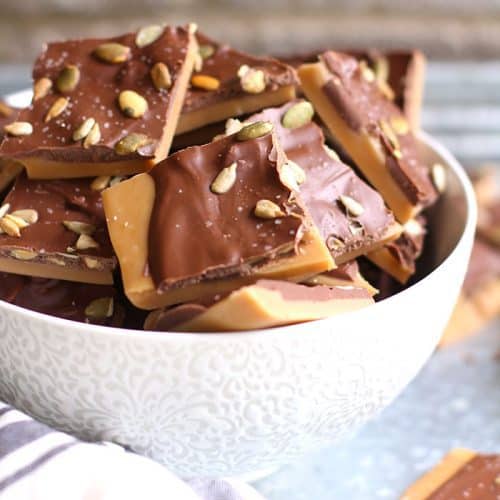 A side shot of a white bowl filled with toffee, on a gray tray with brick wall in the background.