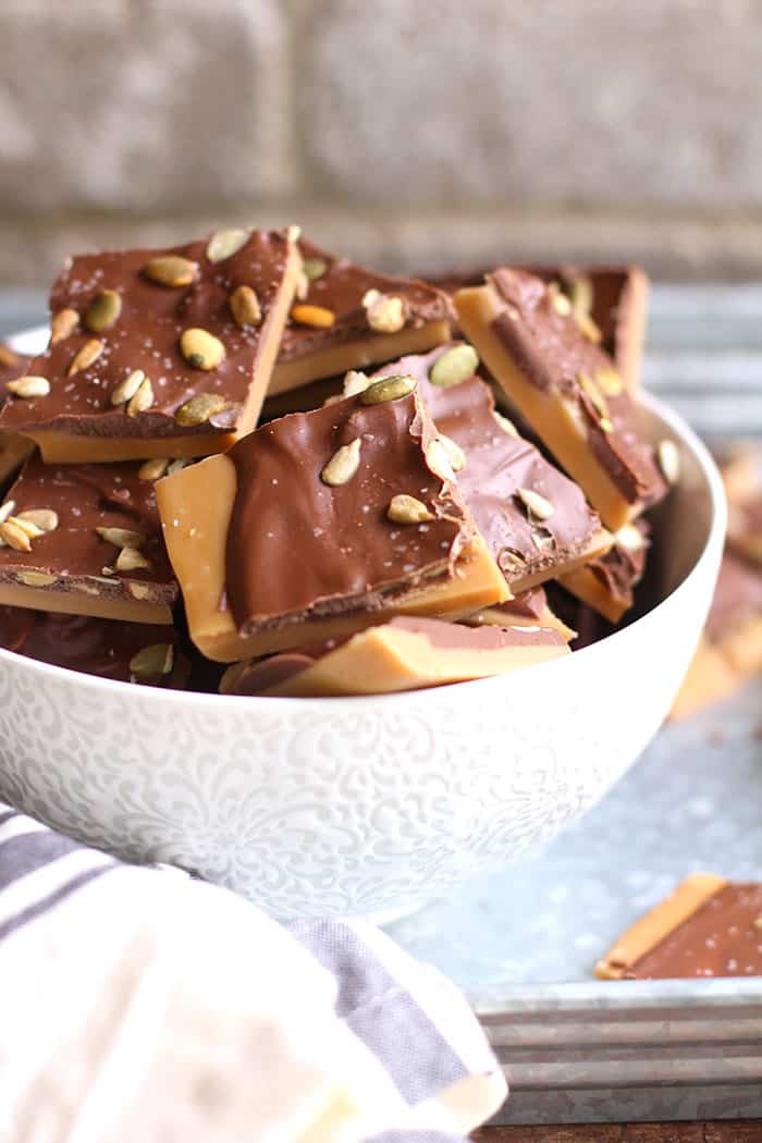 A side shot of a white bowl filled with toffee, on a gray tray with brick wall in the background.