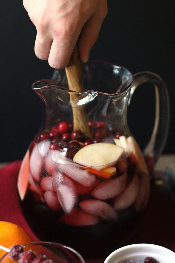 A side shot of a hand holding a wooden spoon, stirring up a large pitcher of holiday cranberry apple sangria, with a black background.
