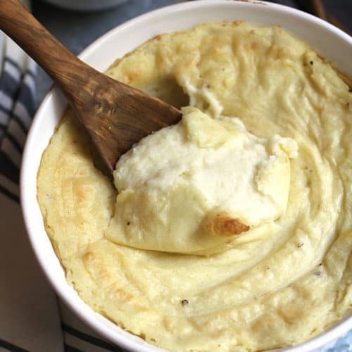 Overhead shot of creamy mashed potatoes in a round white casserole dish, with a wooden spoon in the middle, and you can see how creamy it is, on a gray tray.