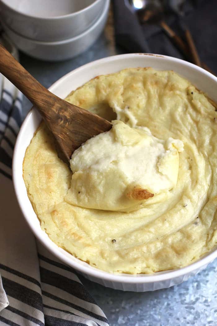 Overhead shot of creamy mashed potatoes in a round white casserole dish, with a wooden spoon in the middle, and you can see how creamy it is, on a gray tray.