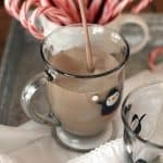A stream of hot chocolate being poured into a glass mug, with candy canes in the background.