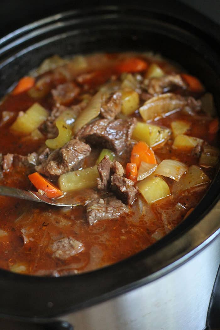 Side shot of a large black slow cooker filled with beef stew in a dark orange broth.