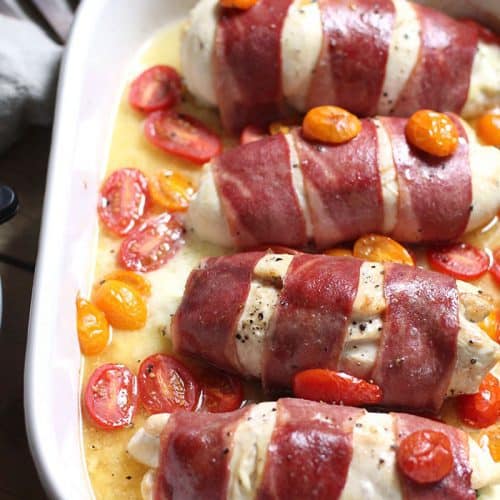 Overhead shot of four turkey wrapped chicken breasts in a white casserole dish with handles, sprinkled with cherry tomatoes.