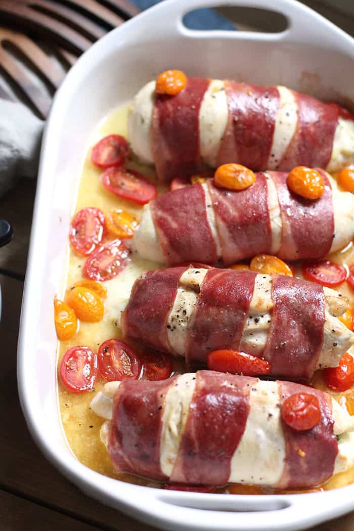 Overhead shot of four turkey wrapped chicken breasts in a white casserole dish with handles, sprinkled with cherry tomatoes.