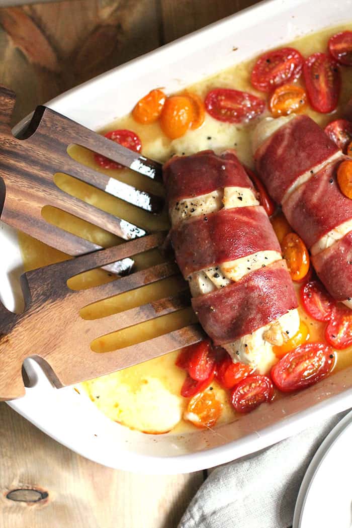 Overhead shot of turkey wrapped chicken breasts, showing two breasts with wooden forks on one side of the white casserole dish.