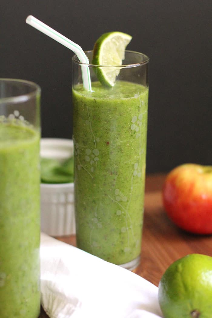 Two glasses of green smoothies, with fruit in the background.