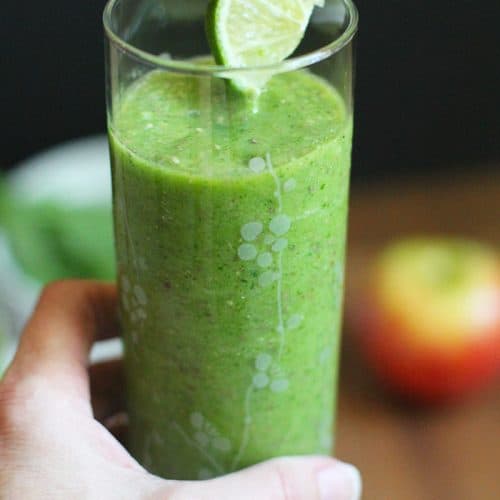 A hand holding a tall glass of ultimate green power smoothie, with a lime wedge.