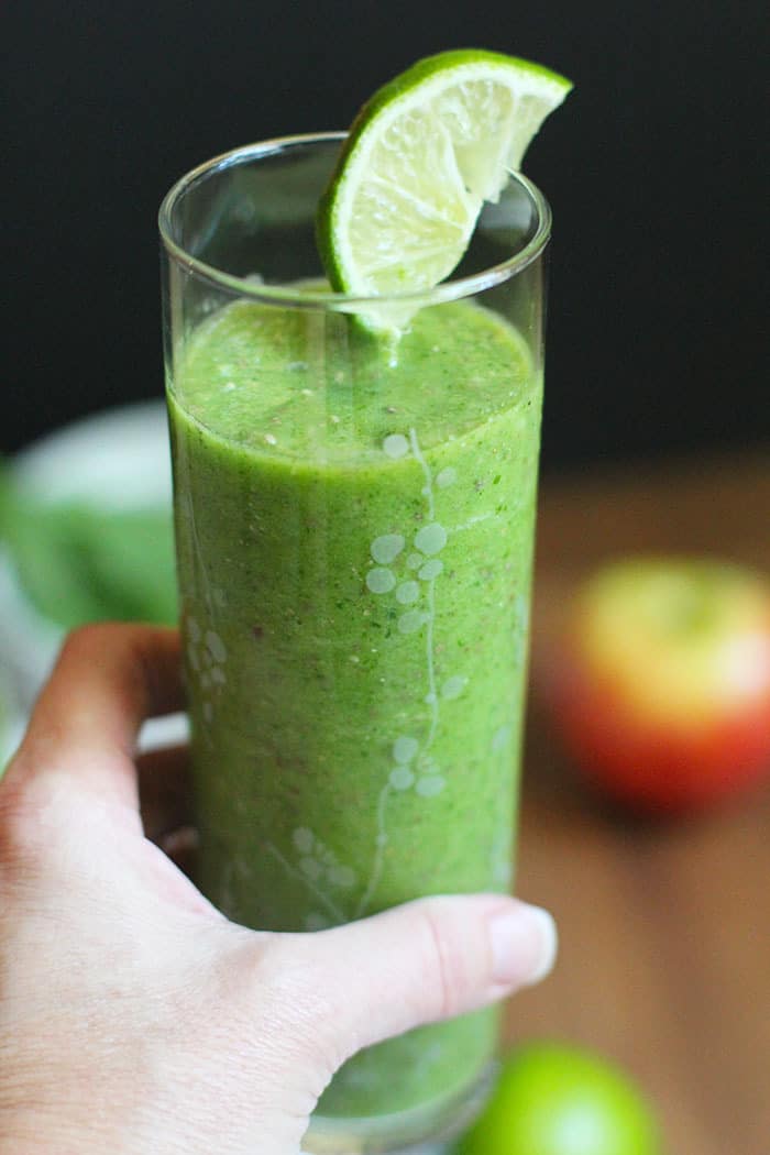 A hand holding a tall glass of ultimate green power smoothie, with a lime wedge.