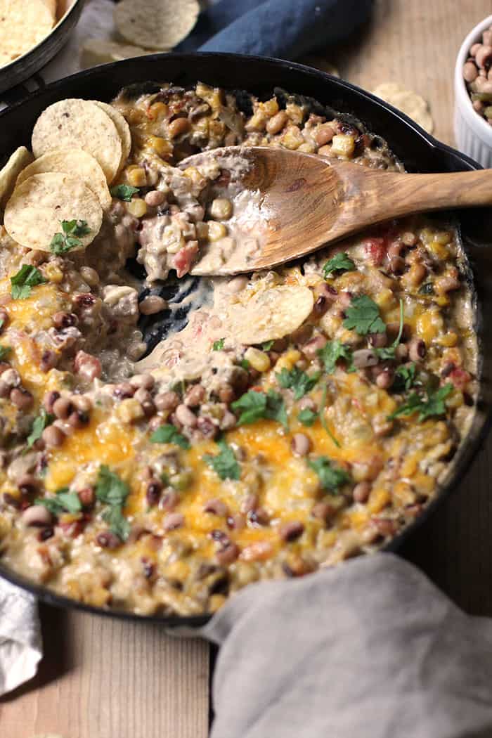 Overhead shot of a cast iron skillet filled with a cheesy black-eyed pea dip, with Tostitos chips and a wooden spoon in the middle of it, on a wooden background.