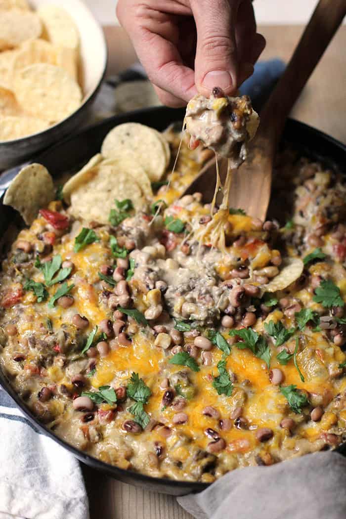 Side shot of a hand picking up a Tostito dipped in the black eyed pea dip, overtop the cast iron skillet of dip.