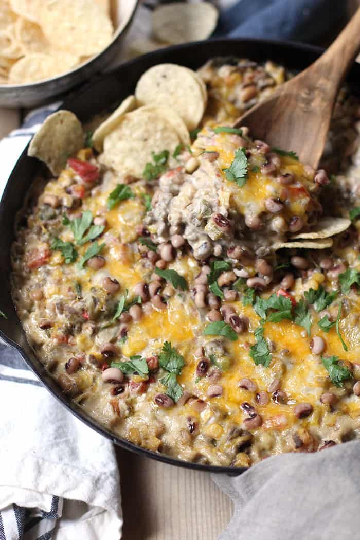 Side shot of a wooden spoon dipping into the cheesy black-eyed pea dip, in a cast iron skillet.