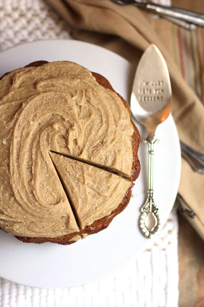 Overhead shot of banana layer cake with caramel frosting, with one piece cut, on a white cake plate with a silver spatula.