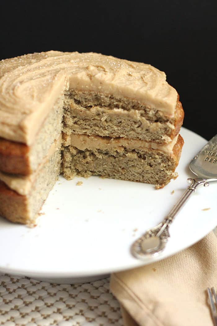 Side angle of banana layer cake with a big slice out of it, so you can see the actual moist layers, all on a white cake plate with black background.