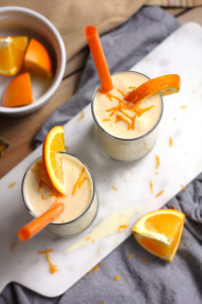 Overhead shot of two glasses of Orange Julius smoothies, garnished with oranges and orange straws, on a white board.