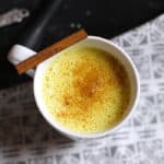 Overhead shot of a white mug of golden milk, with a cinnamon stick onto, on a white and gray napkin.