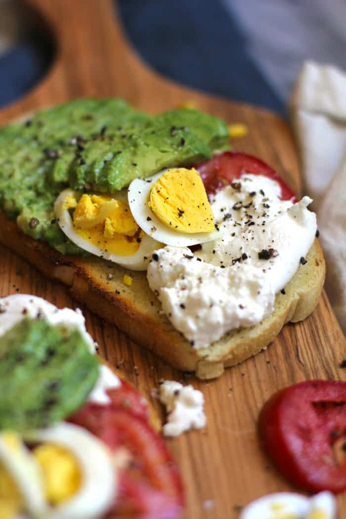 Side view of a piece of toast with burrata cheese, tomato, hard boiled egg, and avocado slices.