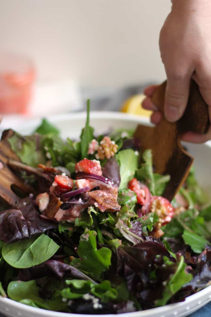 Hands using wooden spoons to serve the strawberry bacon salad.