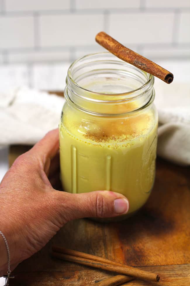 Hand holding jar of anti-inflammatory golden milk, with a cinnamon stick on top.