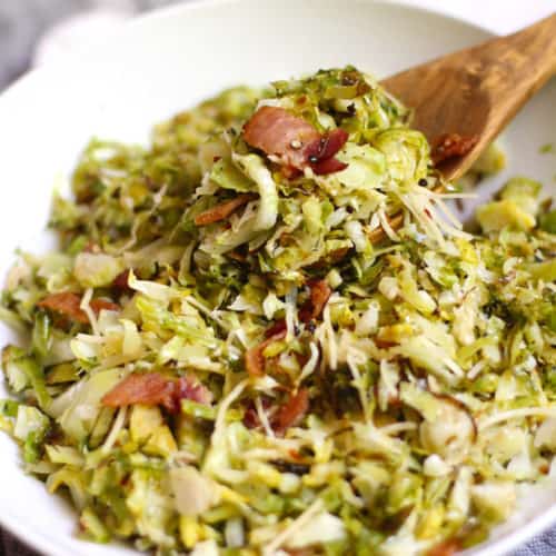 A side shot of a large white bowl filled with shaved Brussels sprouts and bacon, with a wooden spoon scooping some out, on a gray and white background.