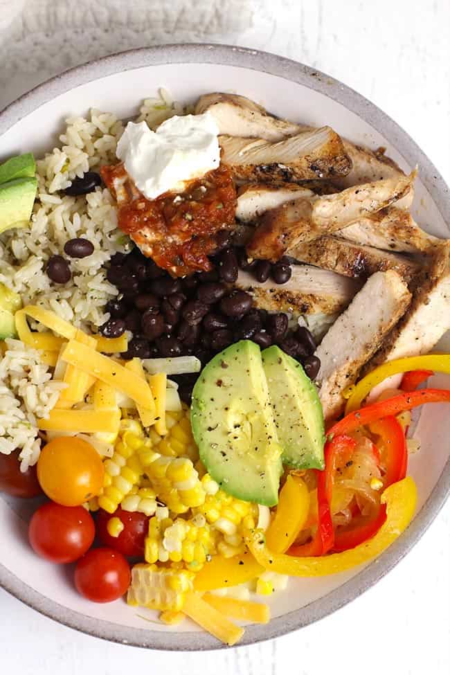 Closeup shot of a white bowl of chicken burrito bowl, with rice, black beans, onions, peppers, corn, tomatoes, cheese, and avocado slices.