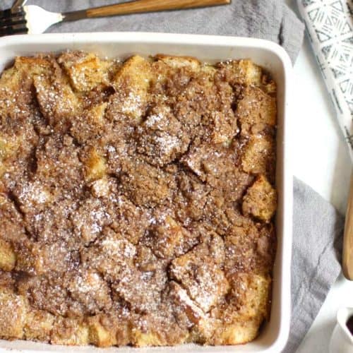 Overhead shot of French toast casserole in a square white dish, on a gray napkin and forks lying nearby.