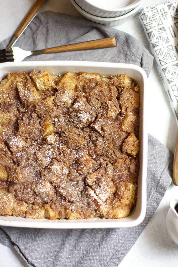 Overhead shot of French toast casserole in a square white dish, on a gray napkin and forks lying nearby.