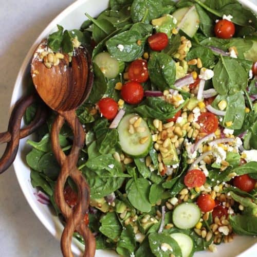 A large bowl of spinach salad with avocado and goat cheese and corn, with a wooden spoon.