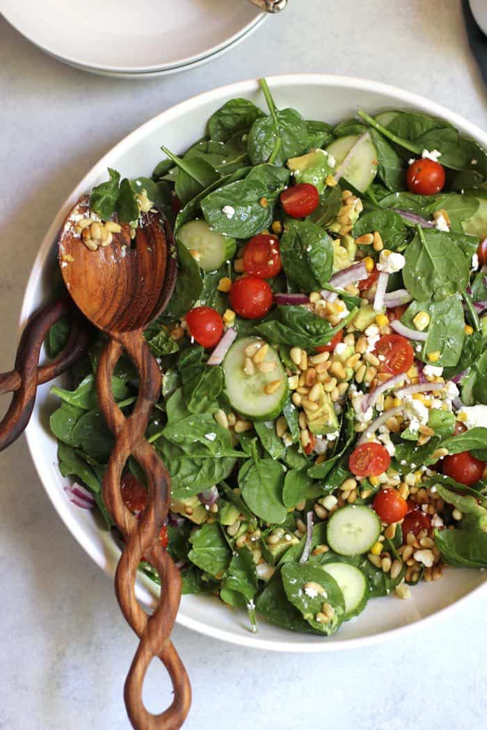 A large bowl of spinach salad with avocado and goat cheese and corn, with a wooden spoon.