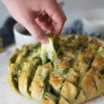 Side view of Cheesy Pesto Party Bread on a white tray, with a hand pulling a pre-cut square and a cheese pull.
