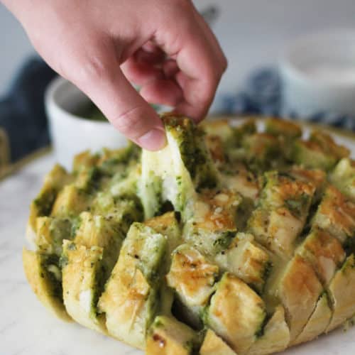 Side view of Cheesy Pesto Party Bread on a white tray, with a hand pulling a pre-cut square and a cheese pull.