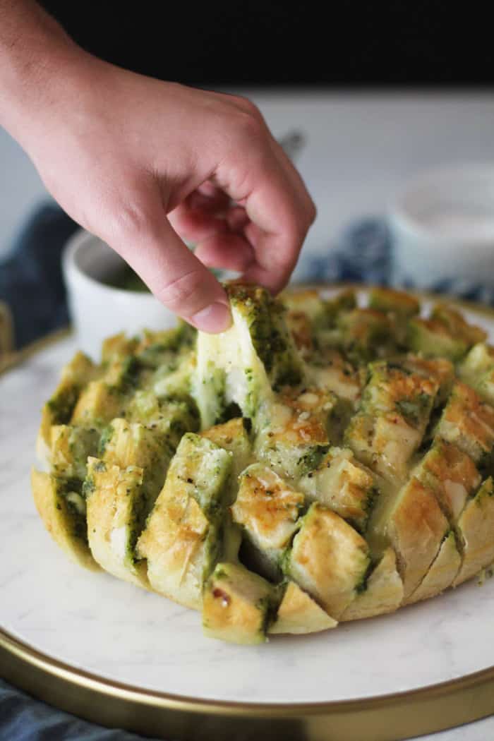 Side view of Cheesy Pesto Party Bread on a white tray, with a hand pulling a pre-cut square and a cheese pull.
