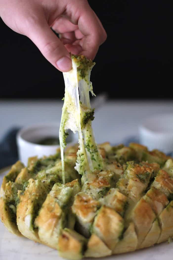 Side view of Cheesy Pesto Party Bread on a white tray, with a hand pulling a pre-cut square and a long cheese pull, amidst a black background.