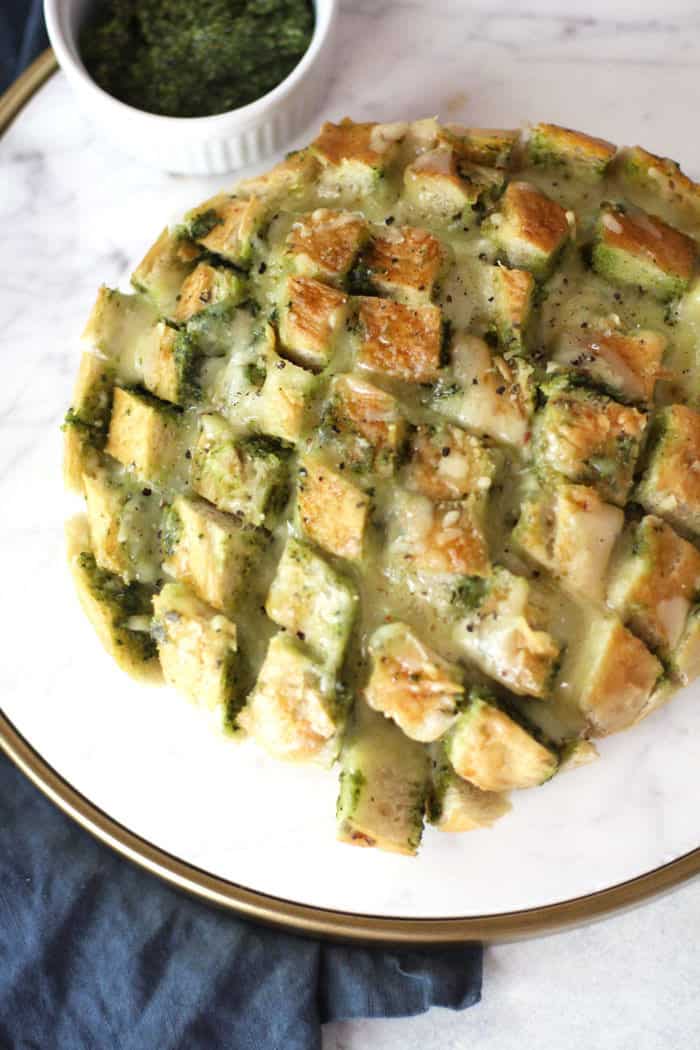 Overhead view of Cheesy Pesto Party Bread on a white tray, with pre-cut squares and an extra bowl of pesto sauce.
