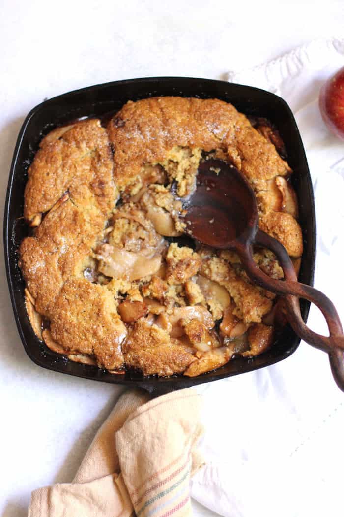 Overhead shot of partially eaten apple cobbler in a square cast iron skillet, with wooden spoon in the center.