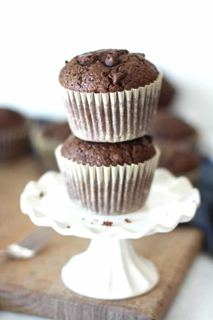 Side shot of two stacked chocolate zucchini muffins on a small white cake plate.