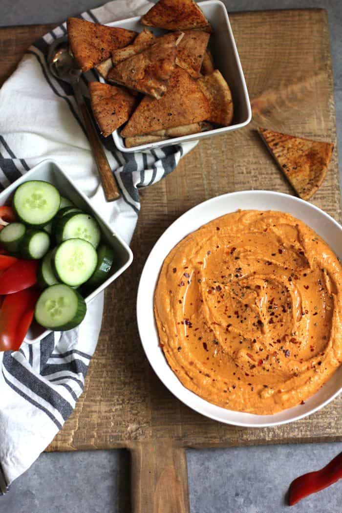 Overhead shot of roasted red pepper hummus in a round white bowl, with pita chips in a square bowl and veggies in a square bowl, all on a wooden cutting board.