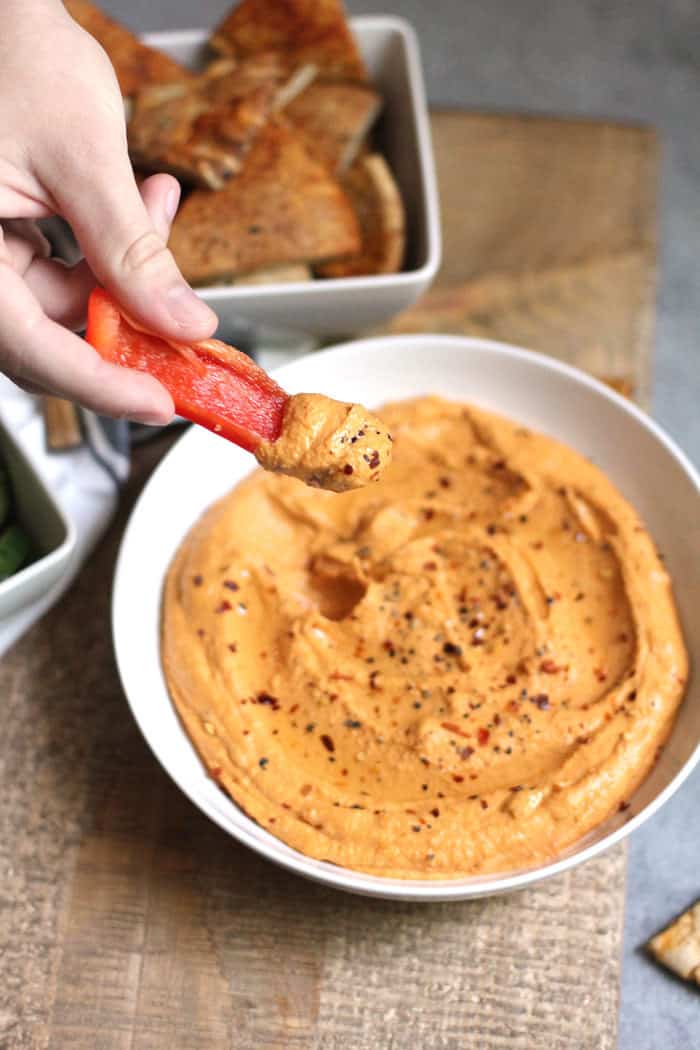 A hand holding a red pepper slice dipped in the hummus.