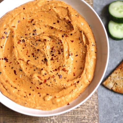 Overhead shot of roasted red pepper hummus in a white bowl, on a brown wooden board, with veggies and pita chips around it.