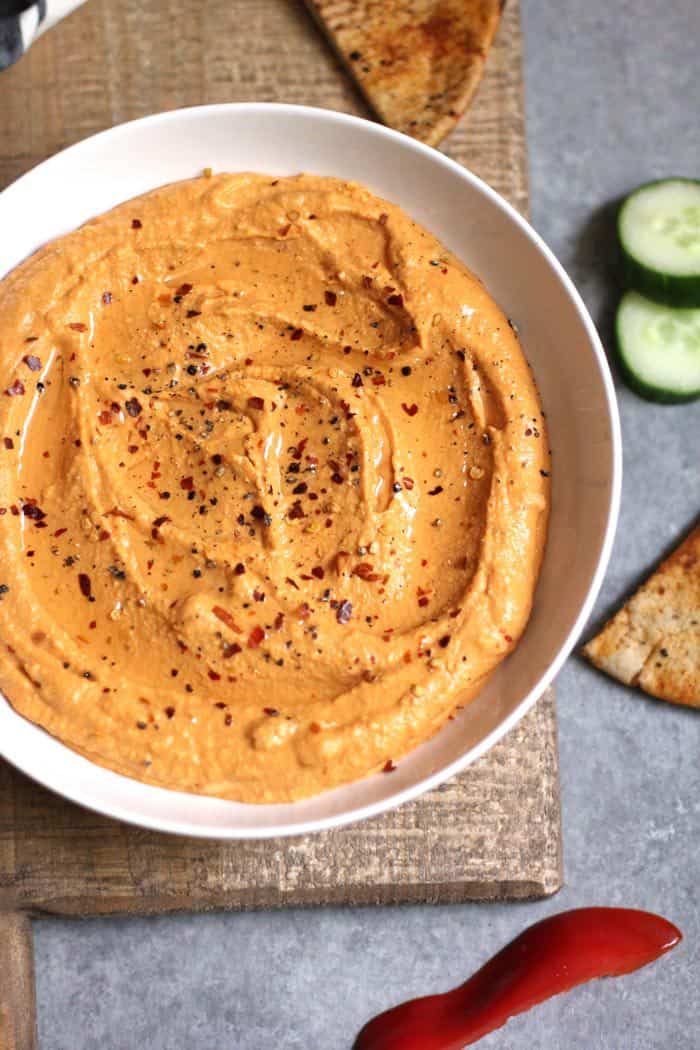 Overhead shot of roasted red pepper hummus in a white bowl, on a brown wooden board, with veggies and pita chips around it.