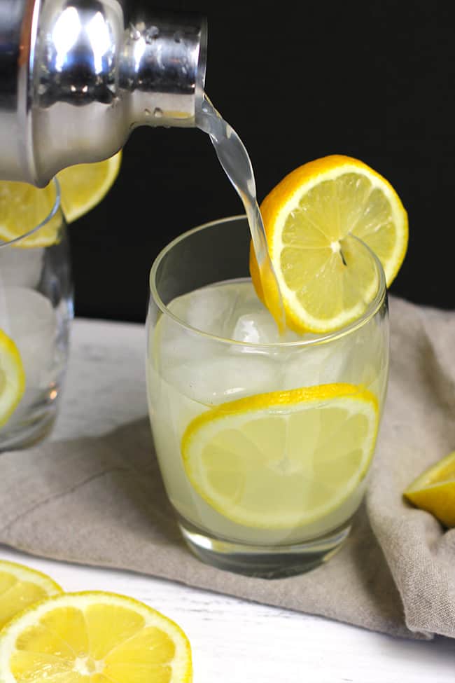 A cocktail shaker pouring cocktails into glasses, with lemon slices.