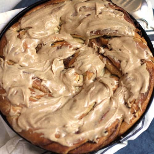Overhead shot of a giant twist cinnamon raisin roll with cinnamon icing in a round cast iron skillet, on a blue napkin.