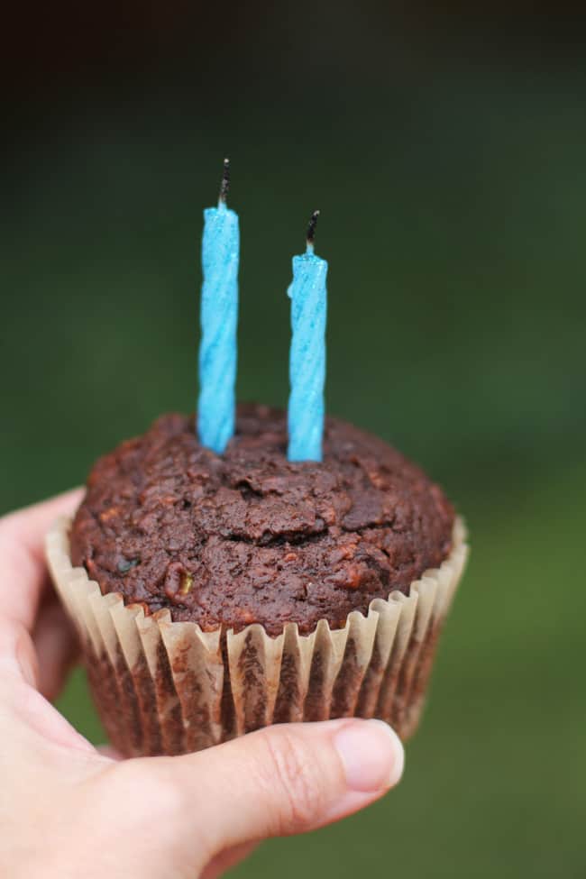 Close-up of my hand holding a chocolate zucchini muffin with two blue candles against a green background.