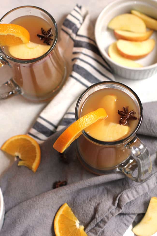 Overhead shot of mulled cider in large clear mugs, with orange slices and apple slices as garnish, on a white background with gray napkins.