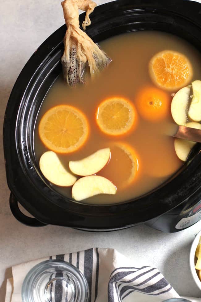 Overhead shot of a large black slow cooker filled with mulled cider, with a cheese cloth full of spices, and orange and apple garnishes.