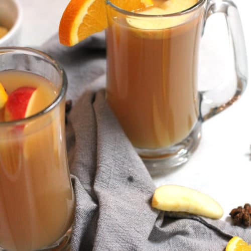 Side angle shot of mulled cider in large clear mugs, with orange slices and apple slices as garnish, on a white background with gray napkins.
