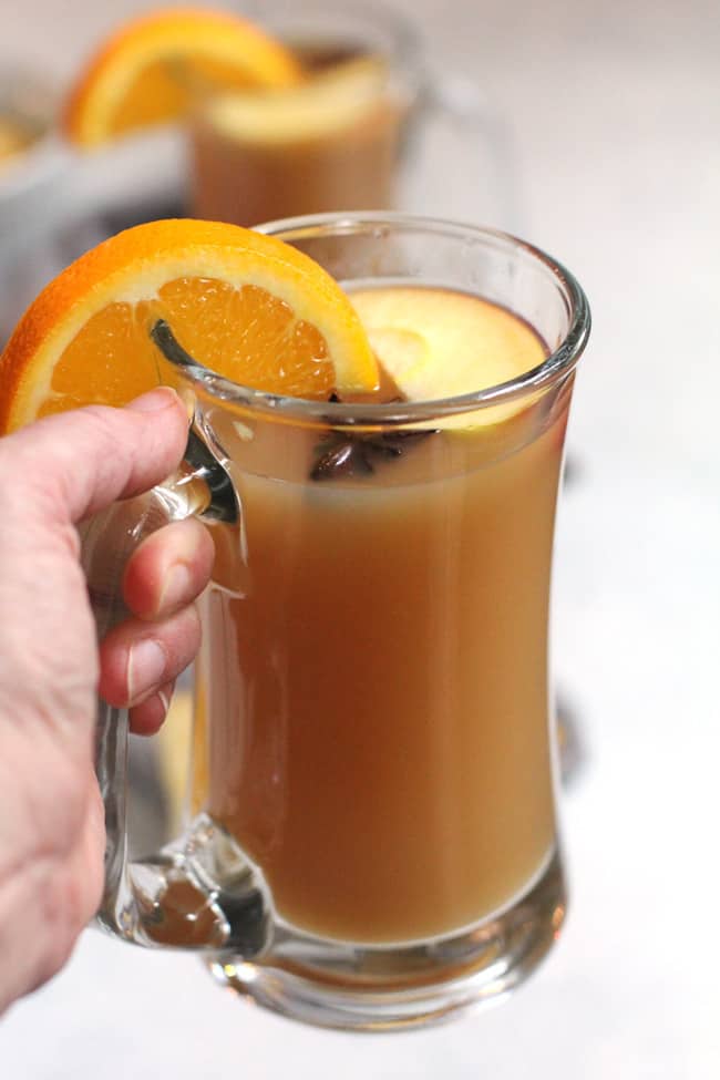 My hand holding a mug of mulled apple cider, with fruit on top.